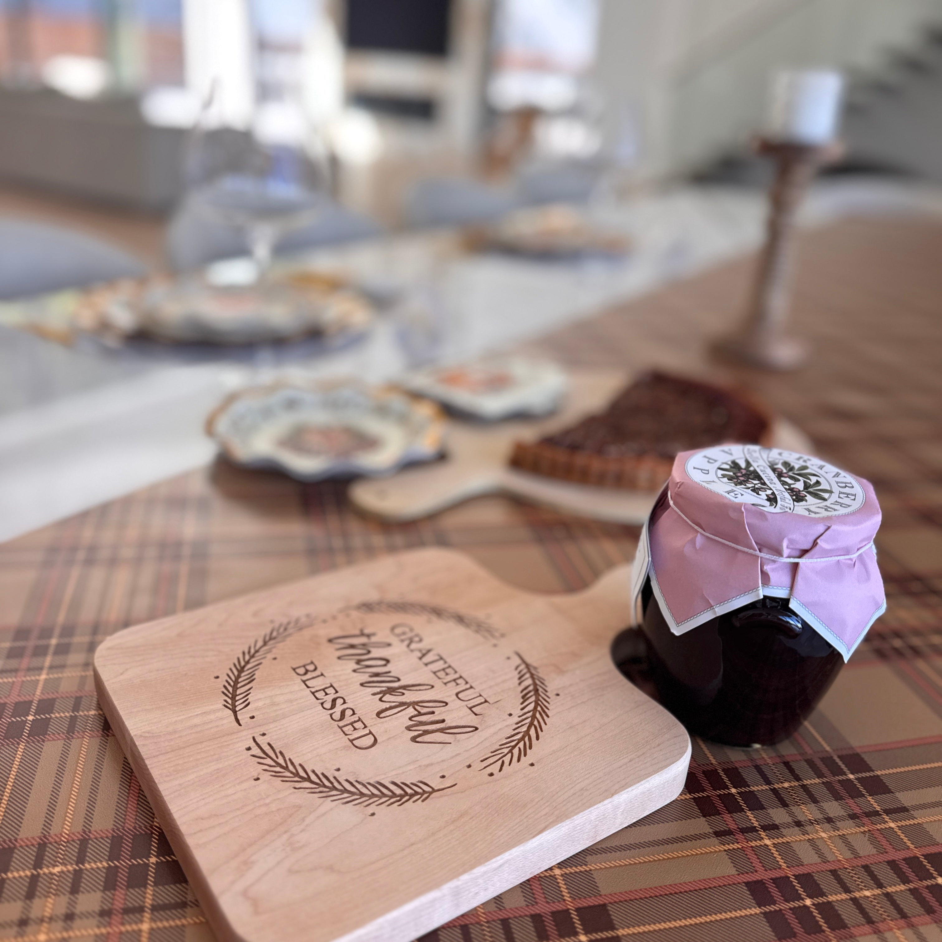 A Turkey & Gourds: Thanksgiving Table for 16 wooden serving board engraved with Grateful, Thankful, Blessed sits on a plaid tablecloth next to a jar of cranberry sauce and a stack of heavy-duty paper plates.