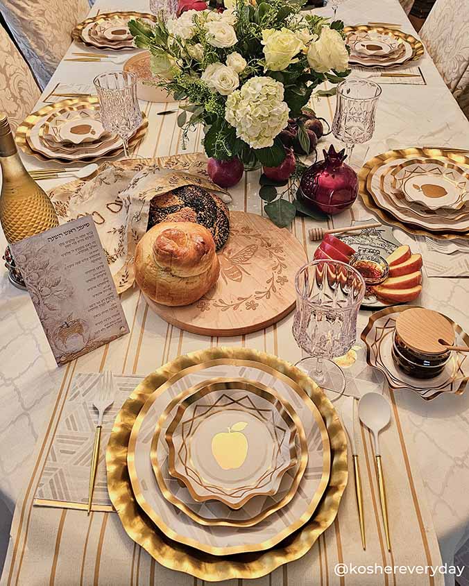A festive table is set for a Jewish holiday with Sophistiplate's Satin Gold Wavy Paper Chargers, wine, challah bread, honey, apples, pomegranates, linen napkins, glasses, and a floral centerpiece for a shimmering celebration.