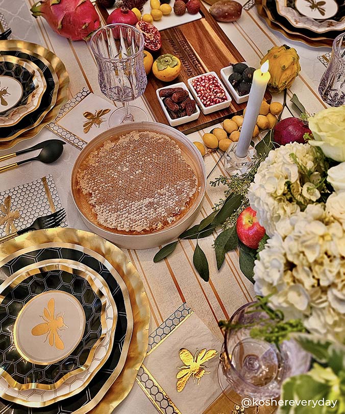A festive table features gold and black bee-themed plates, Sophistiplate Honeybee Paper Cocktail Napkins (26pk), a bowl of honeycomb, candles, flowers, pomegranates, and assorted fruits for an elegant and luxurious atmosphere.