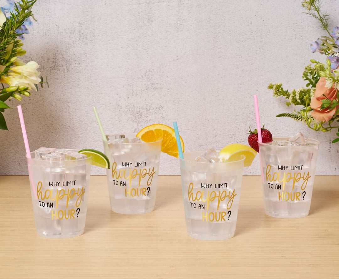 Four clear drinks with ice and fruit garnishes in cups that say Why limit happy to an hour? sit on a table, each with a straw, surrounded by colorful flower arrangements.