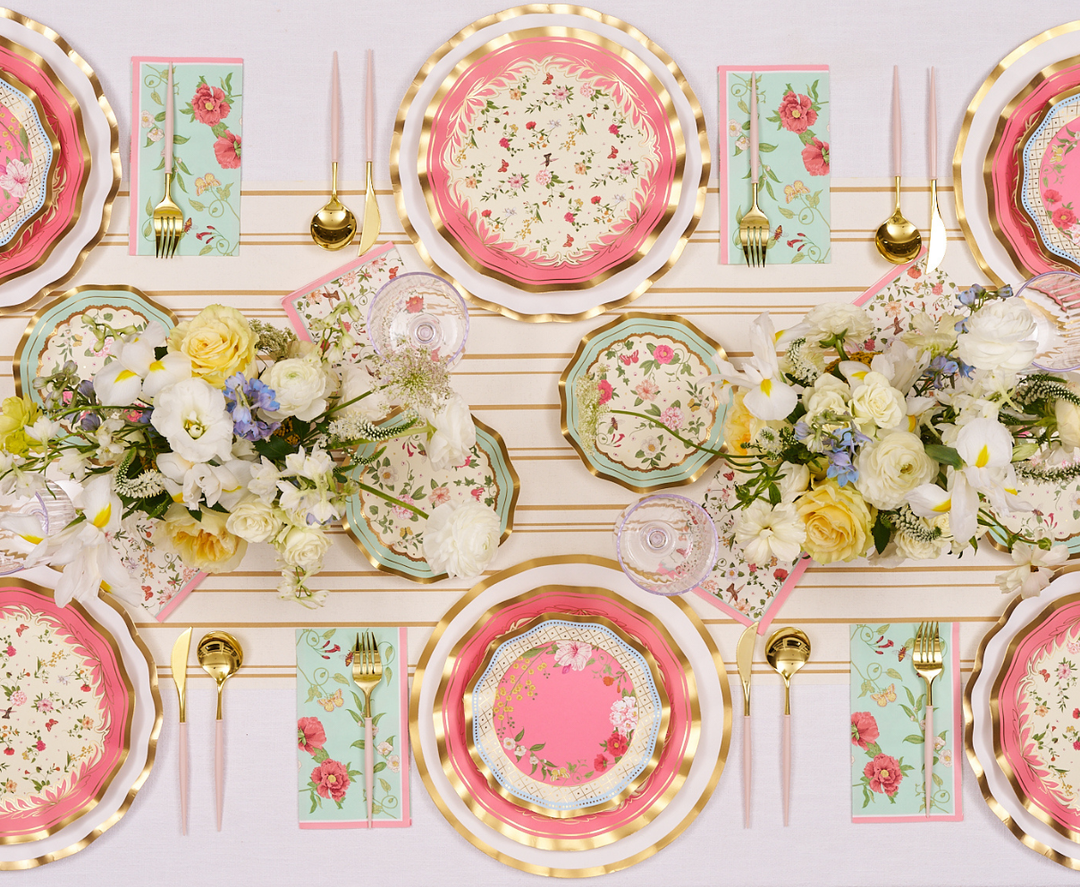 A spring-themed table setting with floral plates, gold cutlery, pastel napkins, pink candles, and white and yellow flower centerpieces arranged on a white tablecloth.