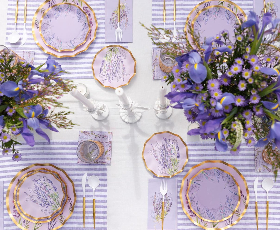 A table set for four with lavender-themed plates, striped placemats, gold utensils, and floral centerpieces of purple flowers. Two white candles are placed in the center.