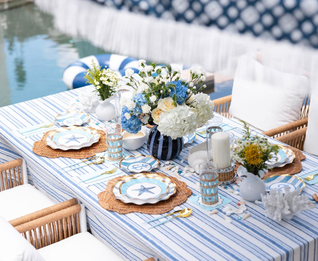 A poolside table decorated with blue and white striped linens, floral arrangements, starfish-themed plates, wicker placemats, and candles, creating a fresh, nautical-themed setting.