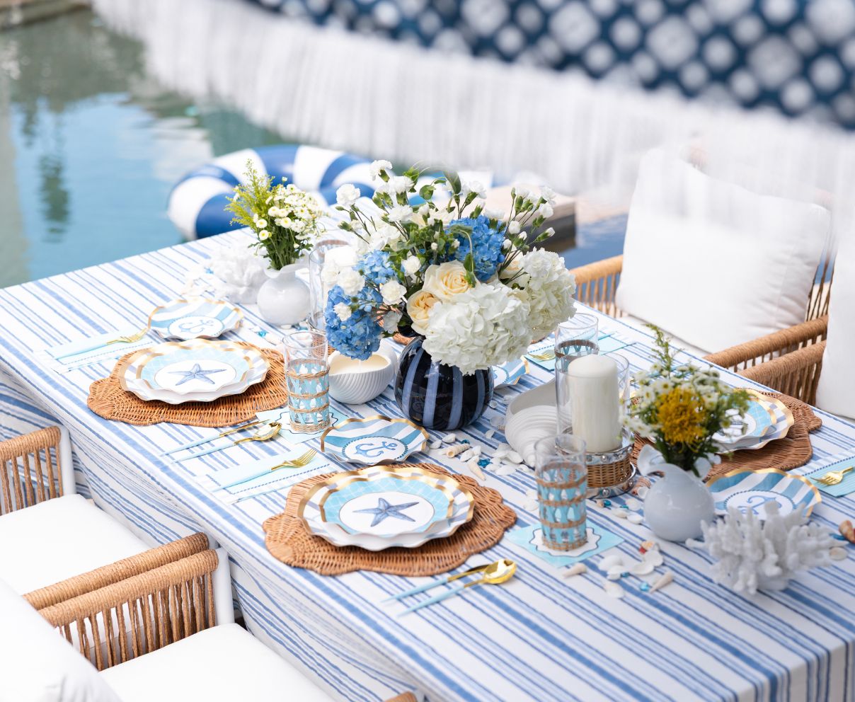 A poolside table decorated with blue and white striped linens, floral arrangements, starfish-themed plates, wicker placemats, and candles, creating a fresh, nautical-themed setting.