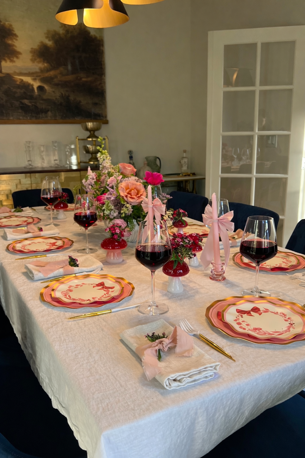 A dining table set for six with pink and gold-rimmed plates, pink candles, wine glasses, gold cutlery, and floral centerpieces in shades of pink and peach on a white tablecloth.