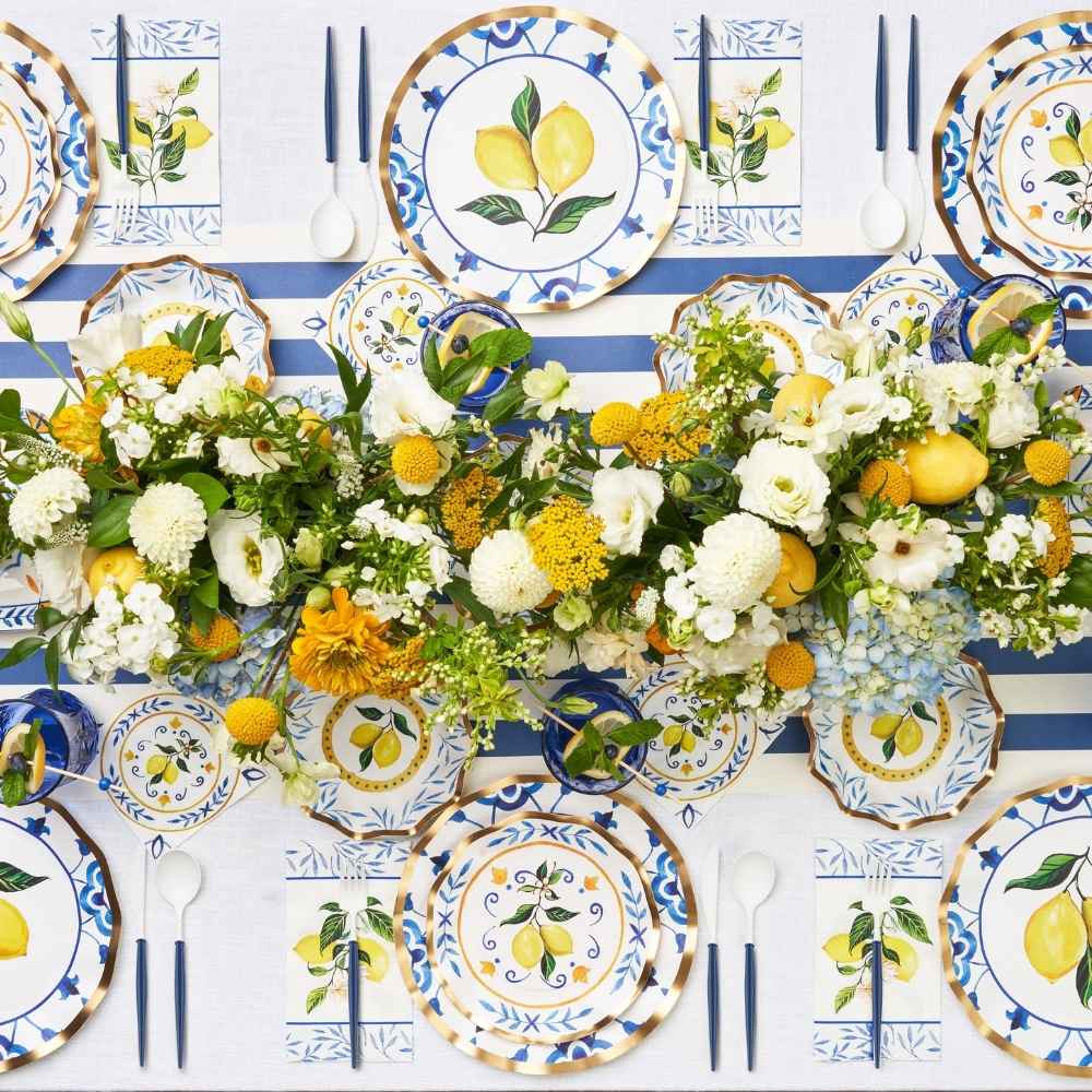 Colorful table setting with lemon-themed plates, blue-striped tablecloth, floral centerpieces with yellow and white flowers, and lemons arranged along the center of the table.