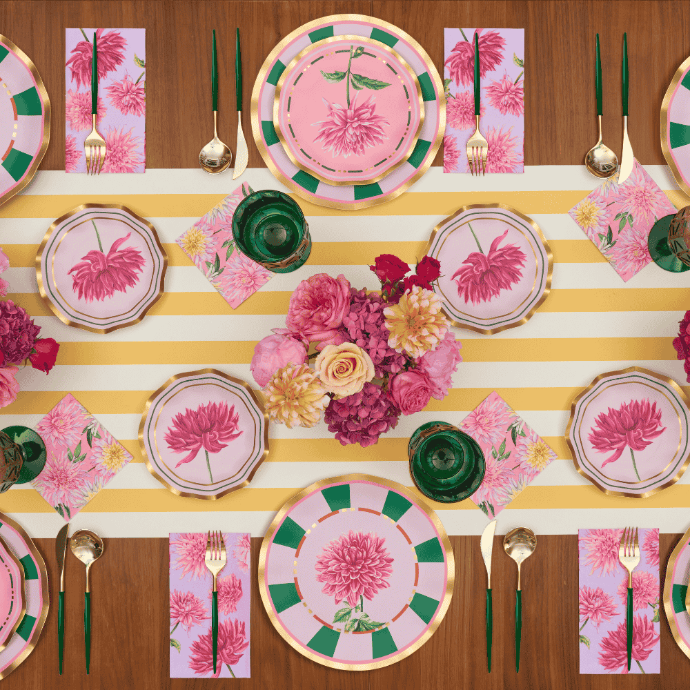 A table set for six with floral plates, patterned napkins, green glassware, and a central bouquet of pink and yellow flowers on a yellow-and-white striped runner.