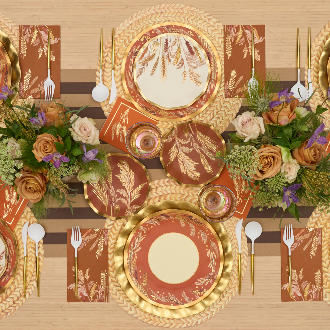 A top view of an elegant table setting with gold and rust-colored plates, floral centerpieces with roses and greenery, gold cutlery, and striped placemats on a wooden table.