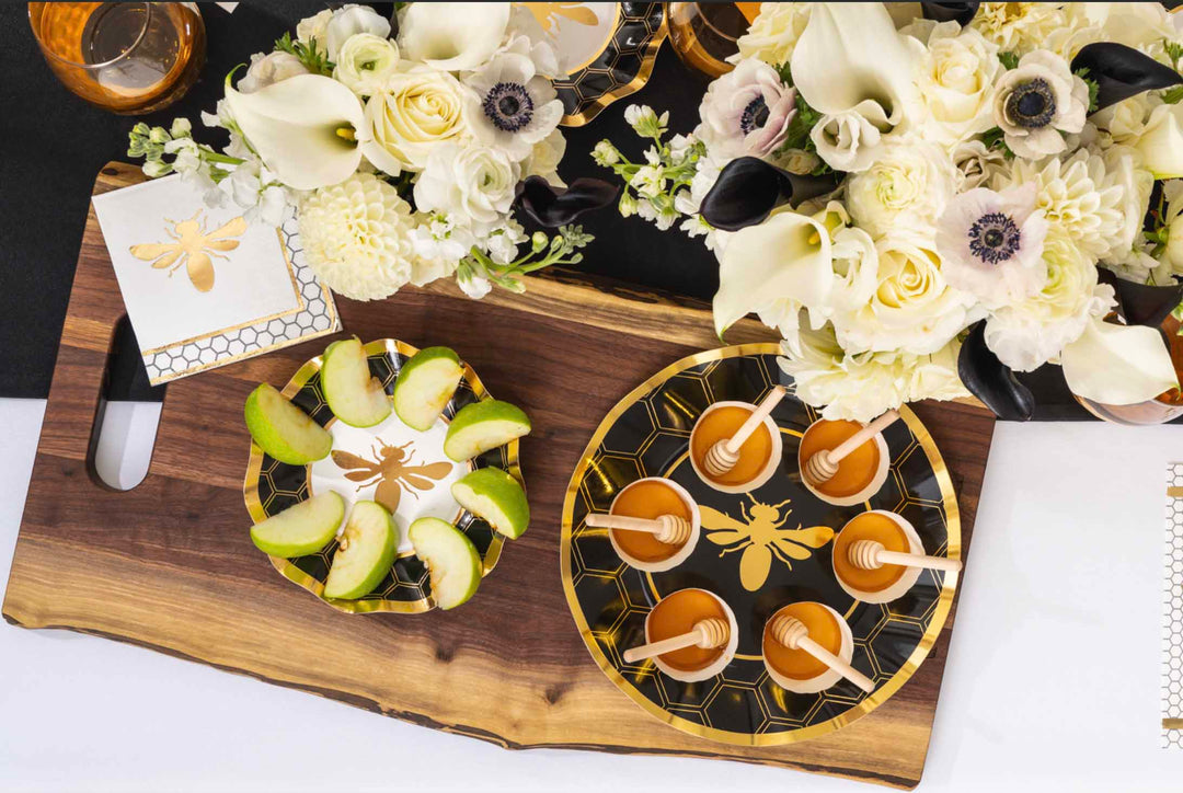 A Rosh Hashanah table set with bee-themed plates, napkins, utensils, a honey jar, and the Maple Bevel Board with Spreader Knife—engraved with “Shana Tova” and a bee graphic (9.75 x 8”)—all arranged on a white patterned tablecloth.