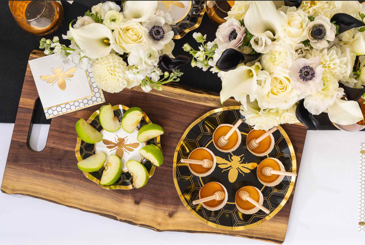 A Rosh Hashanah table set with bee-themed plates, napkins, utensils, a honey jar, and the Maple Bevel Board with Spreader Knife—engraved with “Shana Tova” and a bee graphic (9.75 x 8”)—all arranged on a white patterned tablecloth.