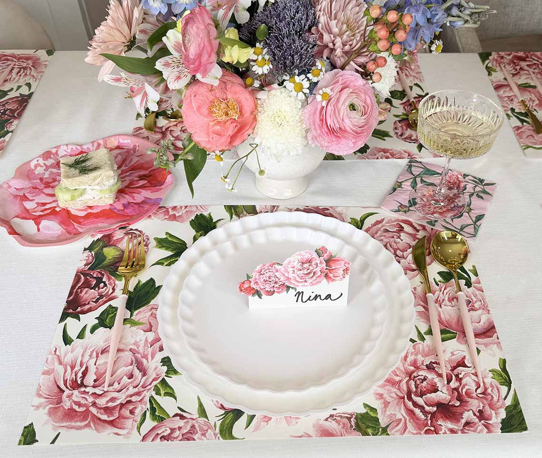 A floral-themed table setting with a white plate, gold cutlery, a glass, and a Nina name card. Pink and white flowers decorate the placemat and centerpiece bouquet.