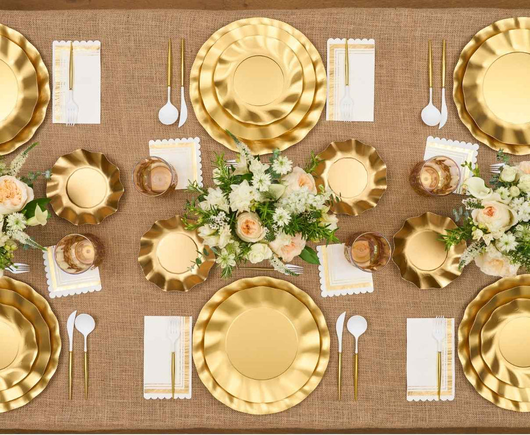A table set for six with gold plates, gold utensils, white napkins, glasses, and flower centerpieces on a burlap tablecloth, arranged symmetrically for a formal meal.