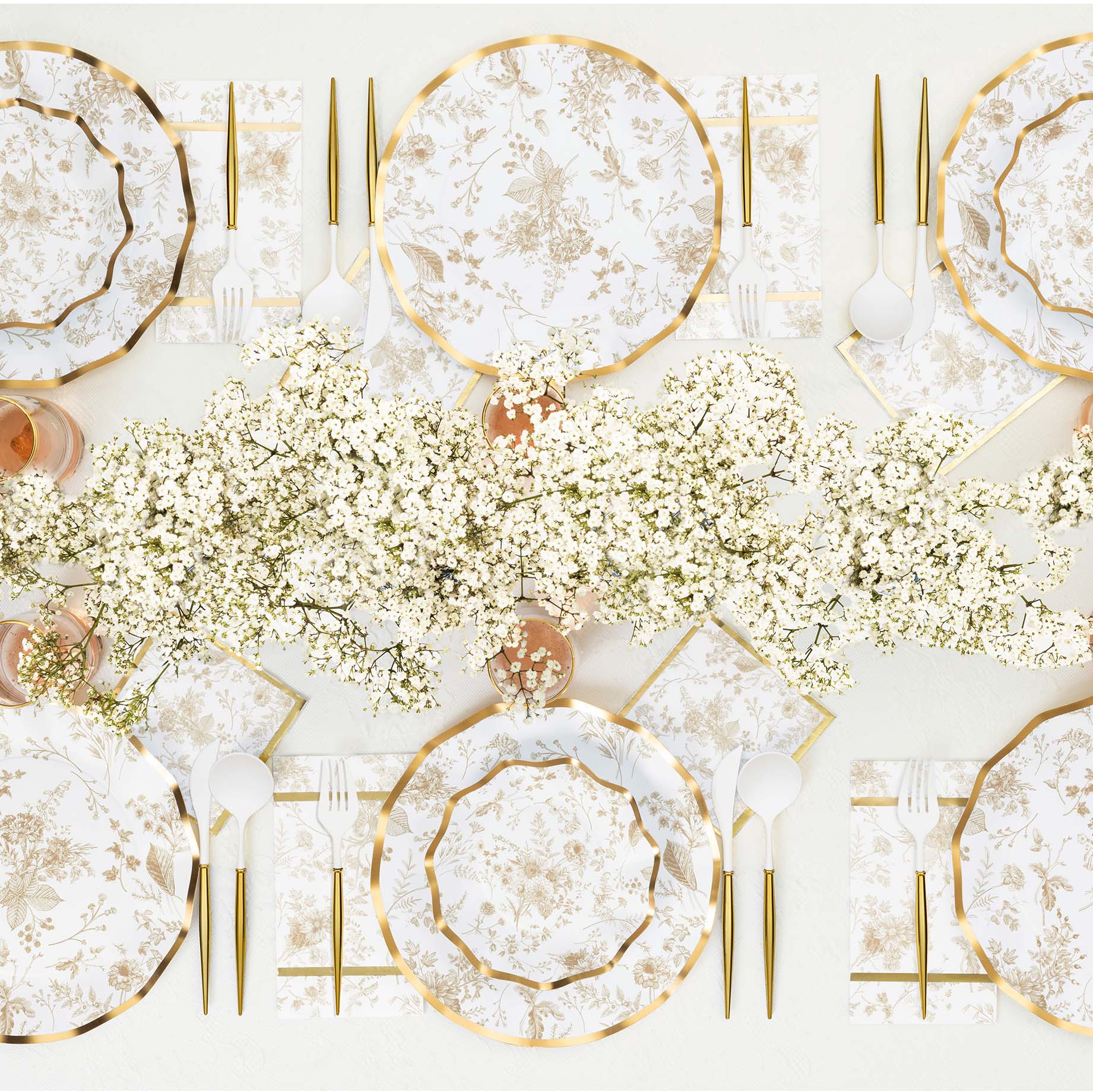 Elegant table setting with gold-rimmed floral plates, gold flatware, and floral napkins. A centerpiece of white babys breath flowers runs along the middle of the table.