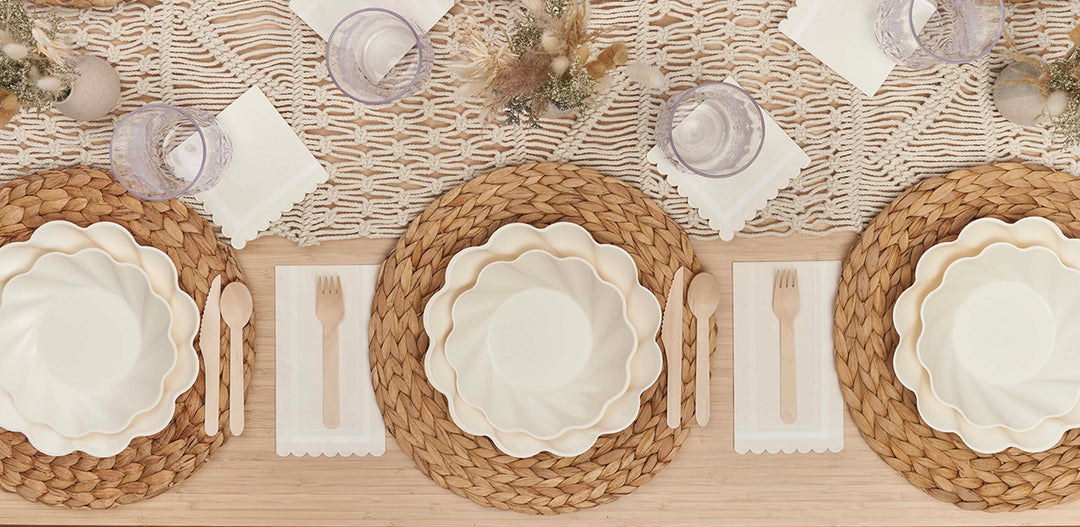 A table set for three with woven placemats, scalloped cream plates, wooden utensils on white napkins, clear drinking glasses, and a textured macrame table runner with dried floral arrangements.