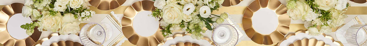 White roses and greenery arranged above shiny gold and white geometric-patterned tableware, including scalloped-edged plates and clear glassware, creating an elegant and festive celebration setting.