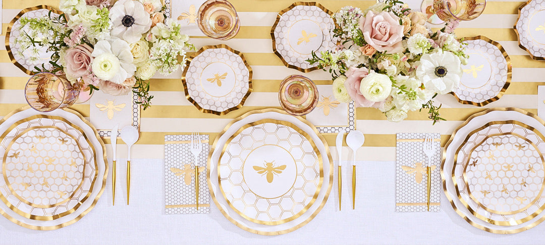 Elegant table setting with gold and white striped tablecloth, gold utensils, floral centerpieces, and plates featuring a bee and honeycomb design. Glasses and napkins arranged neatly.