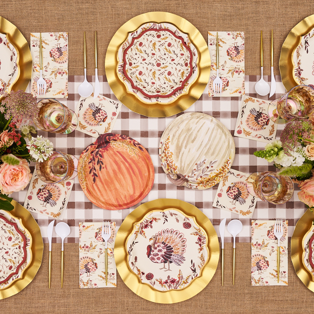 A Thanksgiving table set with floral plates, gold chargers, autumn-themed napkins, gold cutlery, pumpkins, and flower arrangements on a brown checked tablecloth. Sophistiplate’s Amber Harvest Guest Towels (20pk) add a thoughtful touch to each place setting, perfectly complementing the seasonal décor.