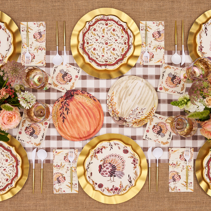 A Thanksgiving table set with floral plates, gold chargers, autumn-themed napkins, gold cutlery, pumpkins, and flower arrangements on a brown checked tablecloth. Sophistiplate’s Amber Harvest Guest Towels (20pk) add a thoughtful touch to each place setting, perfectly complementing the seasonal décor.