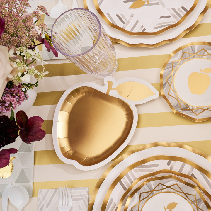 A table set with Sophistiplate's Golden Apple Die Cut Apple Salad Plates, a striped tablecloth, clear ribbed glasses, silverware, napkins, and a floral arrangement featuring pink, white, and burgundy flowers creates an elegant golden apple-inspired setting.