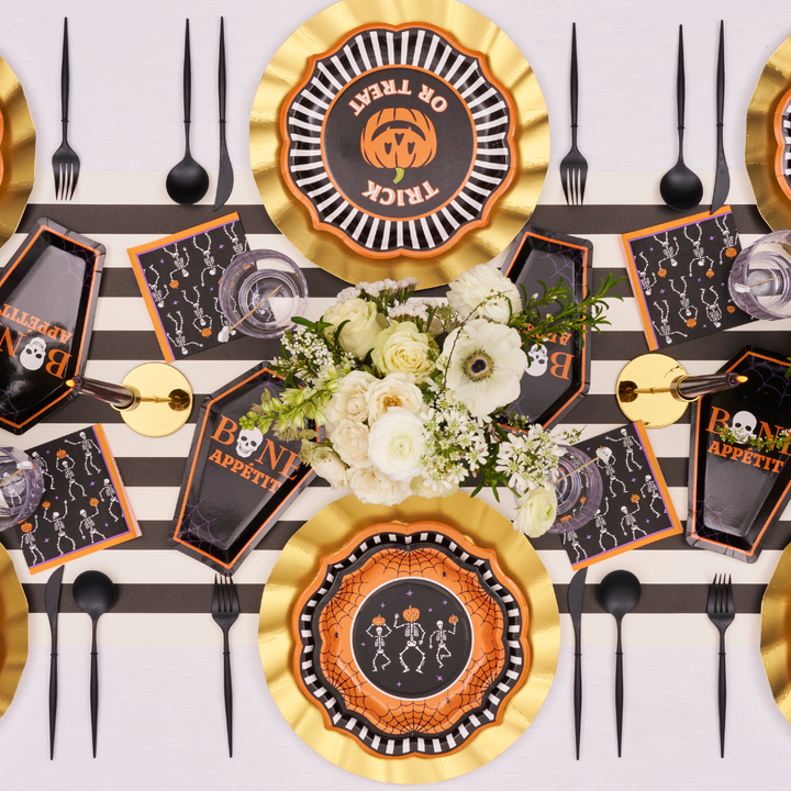 Create a spooktacular Halloween tablescape with a striped tablecloth, gold plates, black cutlery, festive napkins, and the Haunted Halloween Die Cut Salad Plate by Sophistiplate—featuring bold skeleton and spider designs. A centerpiece of fresh white flowers elevates the scene with elegance, making it perfect for showcasing your favorite Halloween party tableware.