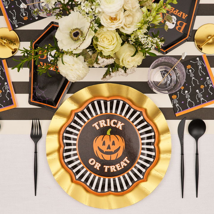 Halloween table setting featuring the Sophistiplate Haunted Halloween Paper Dinner Plate, adorned with black and white stripes and “Trick or Treat” lettering with a jack-o-lantern, complemented by gold accents, black cutlery, skeleton napkins, and a central bouquet of white flowers—a perfect way to highlight spooky tableware from the Sophistiplate Haunted Halloween Collection.