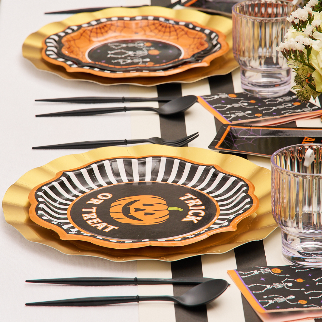 A festive Halloween table setting features gold, black, and orange plates from the Sophistiplate Haunted Halloween Paper Dinner Plate/8pk collection. One plate showcases a jack-o-lantern design with “Trick or Treat” text. The spooky look is finished with black cutlery and clear glasses, all neatly arranged on a white tablecloth.
