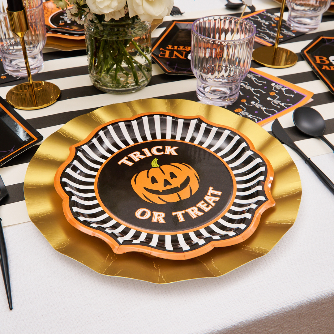 A festive Halloween table setting with a Trick or Treat plate featuring a smiling jack-o-lantern, black and white stripes, gold accents, and black utensils on a striped table runner—perfectly complemented by the Sophistiplate Haunted Halloween Paper Dinner Plate 8-Pack for an extra touch of spooky tableware charm.
