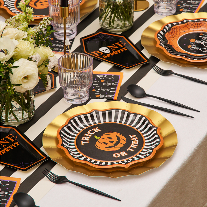 A Halloween-themed table setting from the Haunted Halloween Collection by Sophistiplate features their Haunted Halloween Paper Dinner Plates (8-pack), complemented by black cutlery, clear glasses, bone-printed napkins, and a floral centerpiece—all artfully arranged on a white tablecloth.
