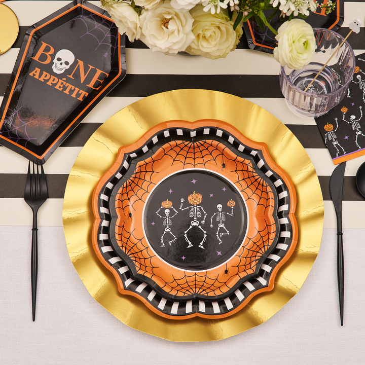 A Halloween-themed table setting featuring the Sophistiplate Haunted Halloween Paper Salad Plate/8pk adds a delightfully eerie touch with skeleton motifs, spiderweb and pumpkin designs, gold and black accents, and a Bone Appetit coffin-shaped plate—all beautifully arranged amidst flowers and utensils for the perfect festive dinner plate display.