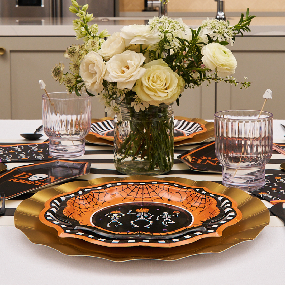 A stylish Halloween table setting featuring the orange and black Haunted Halloween Paper Salad Plates (8pk) from Sophistiplate, paired with gold chargers, elegant glasses, and a floral centerpiece of white blooms arranged in a charming glass jar. Perfect for showcasing your favorite Sophistiplate dinnerware and creating a festive atmosphere.