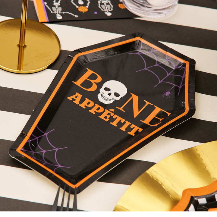 Elevate your Halloween party tableware with the Sophistiplate Haunted Halloween Die Cut Salad Plate/8pk, featuring a unique coffin shape, “BONE APPÉTIT” lettering, and a skull design from the Haunted Halloween Collection—perfectly displayed on a black-and-white striped table and surrounded by gold and spooky decorations.