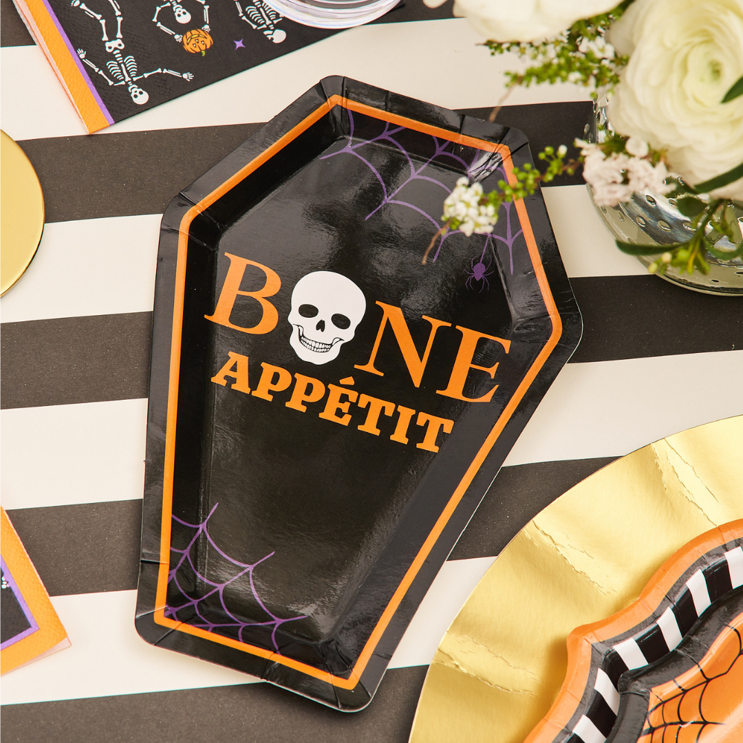 The Haunted Halloween Die Cut Salad Plate/8pk by Sophistiplate features a coffin-shaped black design adorned with a skull, cobwebs, and the phrase “BONE APPÉTIT” in vibrant orange and white. Surrounded by festive Halloween-themed tableware and florals, this plate is a perfect addition to your spooky party spread.