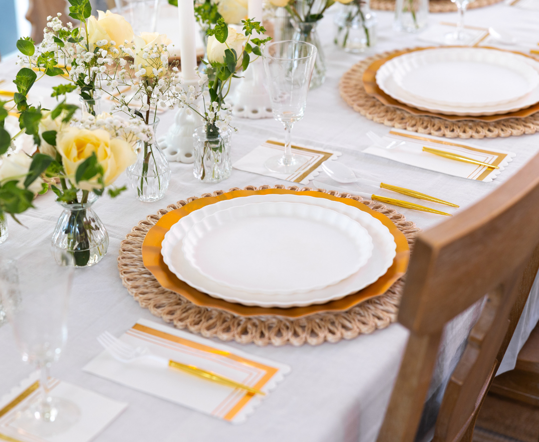 A close-up of the Eco White & Gold Table Setting by Sophistiplate LLC features designer disposable plates, gold and white cutlery, glassware, and vases of yellow roses with greenery on woven placemats atop a white tablecloth.