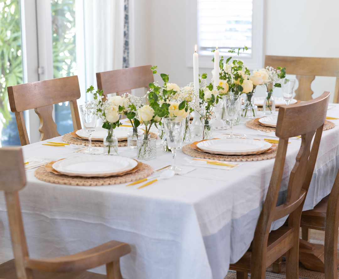 The Eco White & Gold Table Setting by Sophistiplate LLC features party plates on wicker chargers, yellow-handled cutlery, glasses, and wooden chairs for six. The white tablecloth is adorned with white flowers, greenery, and two lit candles.