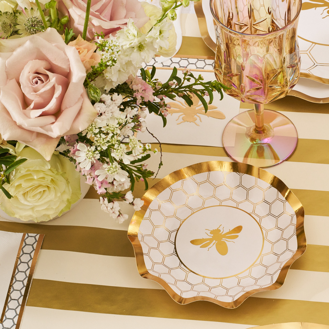 A Honeybee White Wavy Paper Appetizer/Dessert Bowl by Sophistiplate, featuring a gold honeycomb pattern and central gold bee, rests on a striped gold and white tablecloth beside a pink-and-gold glass and roses with greenery.