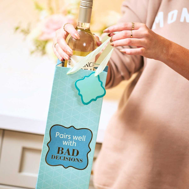 A person places a bottle of white wine into the Drinks On Me Bad Decisions- Wine Bag, featuring bold black and white "Pairs well with BAD DECISIONS" text. Only their hands and torso are visible—perfect as a unique hostess gift.