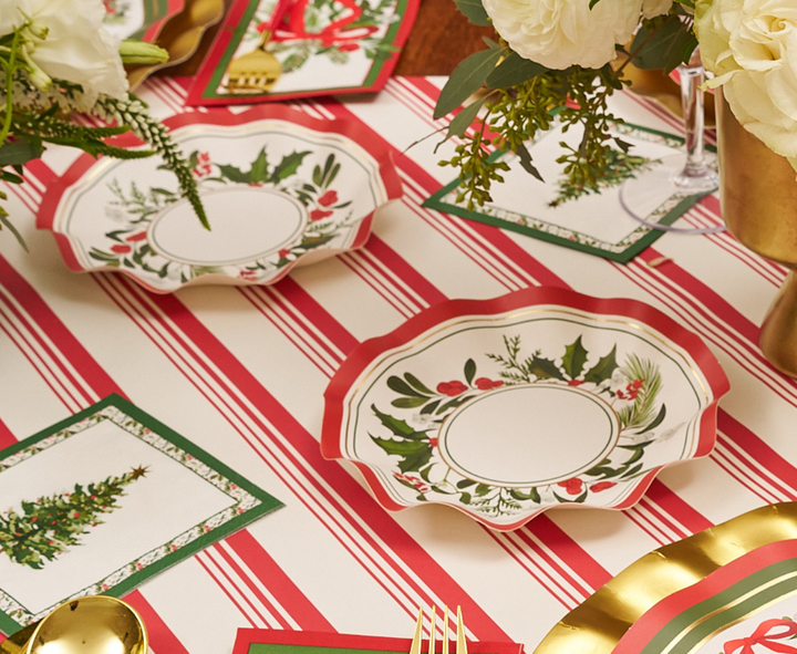 Create a festive table setting with a red and white striped tablecloth, floral plates, napkins featuring a Christmas tree motif, gold cutlery, Sophistiplate’s Home for the Holidays Appetizer/Dessert Bowls (8pk) with elegant wavy edges, and white floral arrangements displayed in gold vases.