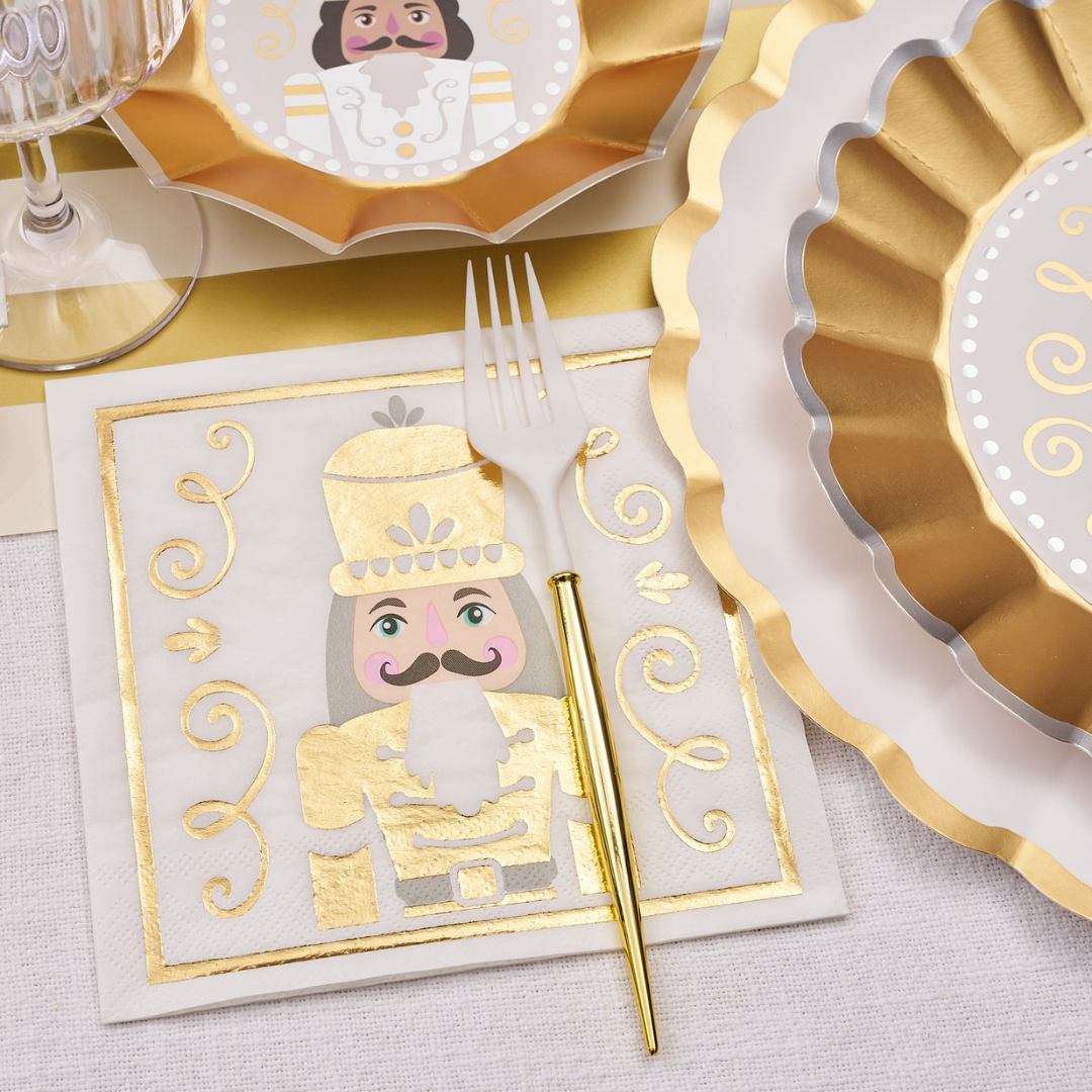The Gilded Nutcracker Table Setting by Sophistiplate features gold and white scalloped plates, a clear wine glass, a festive nutcracker napkin, and a white plastic fork with a gold handle for elegant disposable holiday dining.