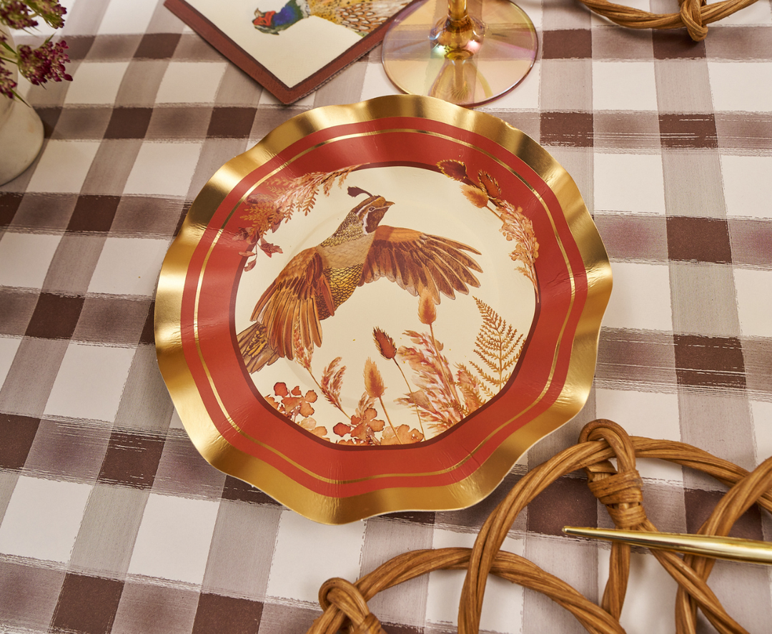 A decorative plate with gold edges and a bird illustration in the center from the Woodland Harvest collection sits on a checkered tablecloth, surrounded by Sophistiplate Woodland Harvest Appetizer/Dessert Bowls (8 pack), elegant table settings, and floral accents.
