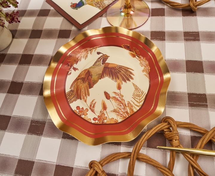 A decorative plate with gold edges and a bird illustration in the center from the Woodland Harvest collection sits on a checkered tablecloth, surrounded by Sophistiplate Woodland Harvest Appetizer/Dessert Bowls (8 pack), elegant table settings, and floral accents.