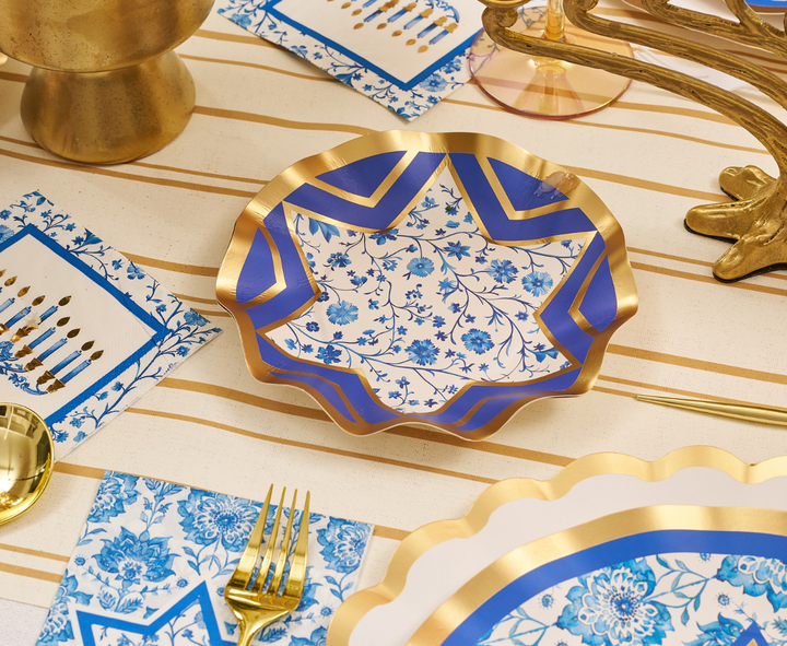 The Hanukkah Toile Table Setting by Sophistiplate features elegant gold and blue floral plates inspired by Hanukkah, complemented by a gold fork and spoon, matching floral napkins, and a striped tablecloth, all accented with coordinating gold-toned serveware.