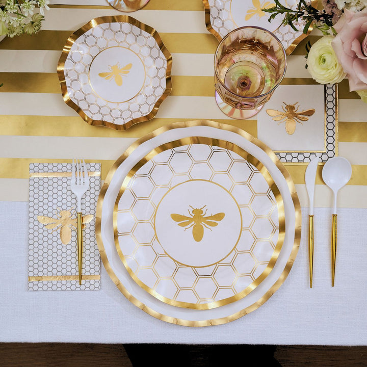 Create a stunning tablescape for any stylish event with the Sophistiplate White Honeybee Table Setting, featuring elegant gold bee and honeycomb designs on gold-rimmed paper plates, matching napkins and cups, coordinating gold and white utensils, and a striped gold and white table runner accented by soft pink flowers in the corner—perfect for elevating your party with beautiful disposable plates.