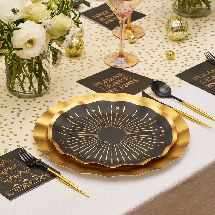 Black square Midnight Sparkle Paper Cocktail Napkin Please Leave By 12:01 /26pk printed with gold text, displayed on a place setting with a gold-rimmed plate and floral centerpiece.