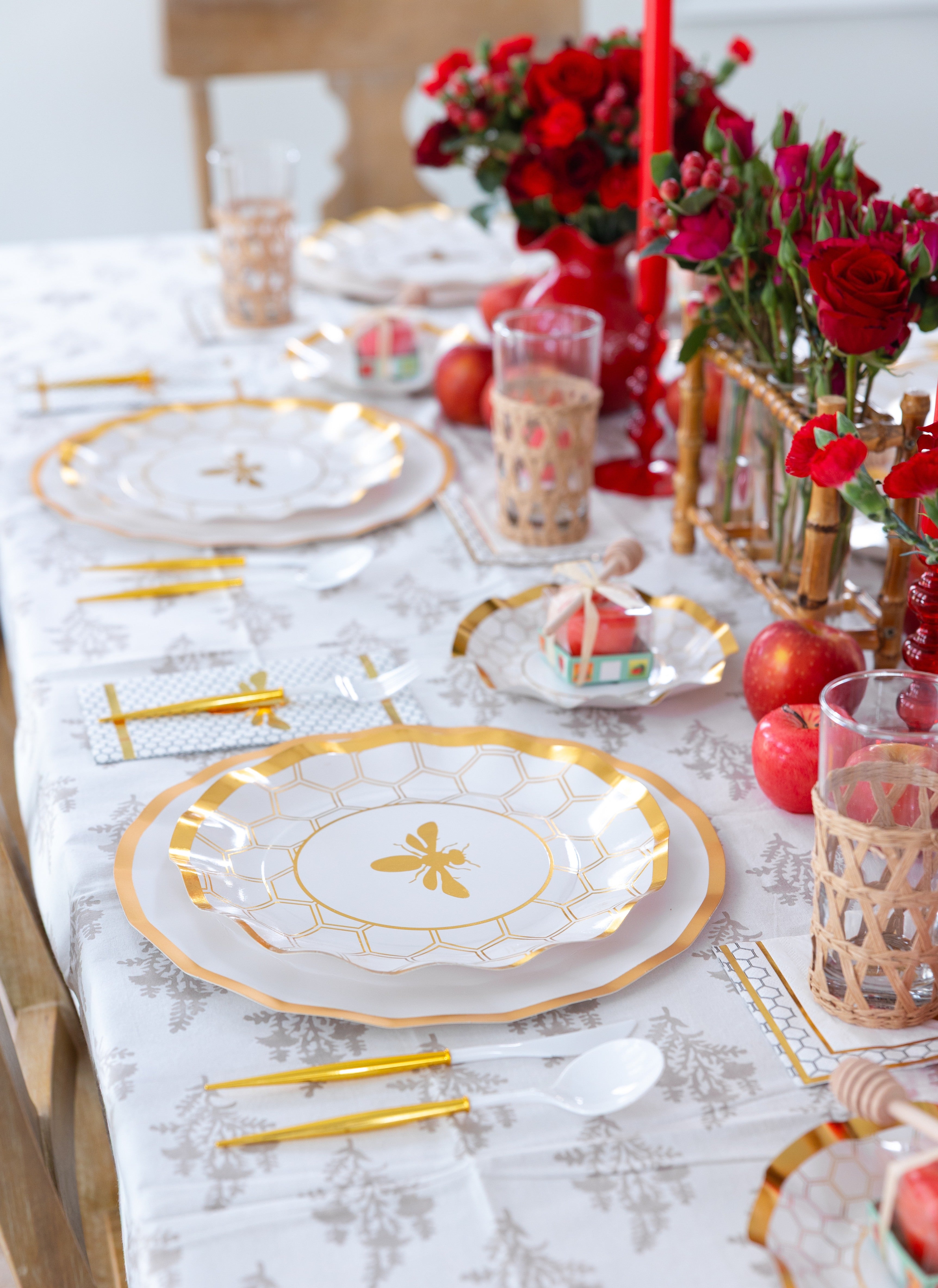 A festive table is set for Rosh Hashanah with the Sophistiplate LLC “Rosh Tablescape for 16 Guests: White Honeybee,” featuring elegant white and gold designer paper plates, gold cutlery, woven glasses, red candles, apples, and vibrant red flowers in vases to create a cheerful and sophisticated celebration.