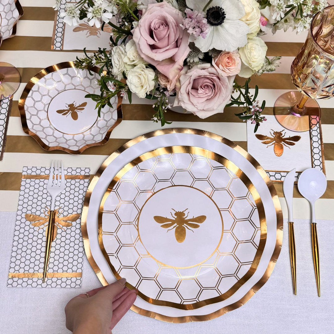 A hand holds the Sophistiplate White Honeybee Table Setting, featuring a gold and white plate with a bee design, surrounded by coordinating gold-rimmed paper plates, napkin, utensils, and a floral centerpiece atop a gold-striped tablecloth.