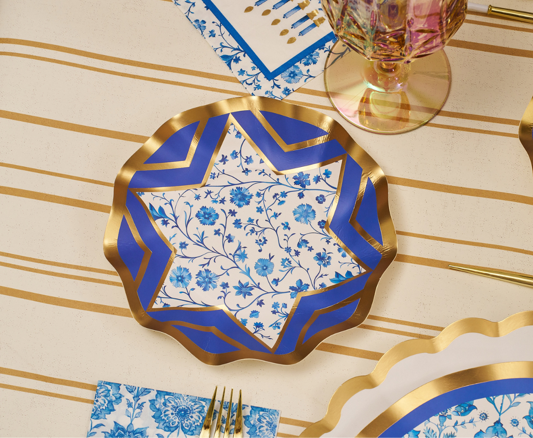 The Sophistiplate Hanukkah Toile Table Setting features a decorative designer paper plate adorned with gold, blue, and white floral patterns on a striped tablecloth. Complementing the plate are gold utensils, a pink glass, and coordinating blue floral napkins, creating an elegant and festive tablescape.