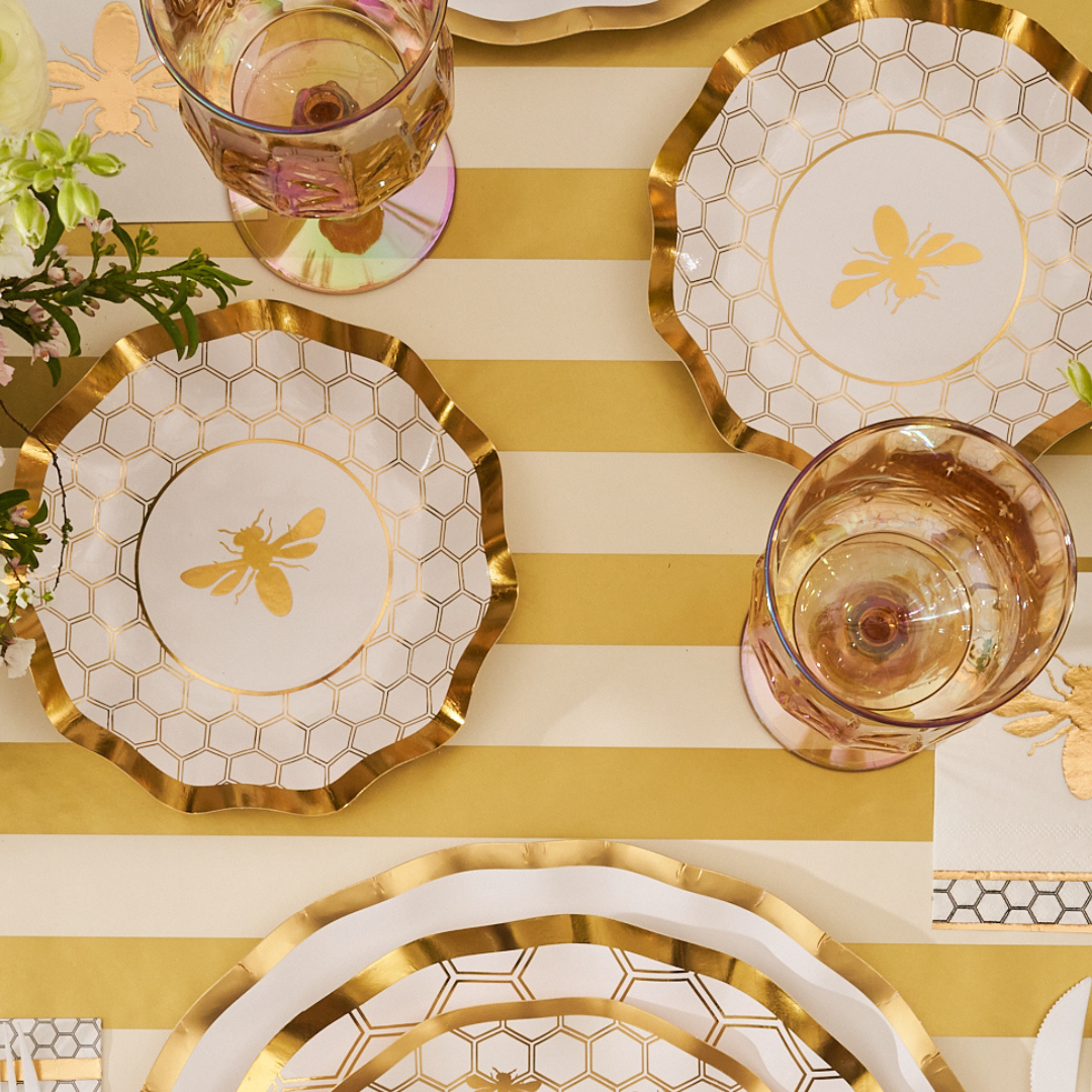 A table is set for a meal with Sophistiplate’s Honeybee White Wavy Paper Appetizer/Dessert Bowls, gold-striped placemats, pink-tinted glasses, gold-edged plates, and white flowers in the corner.