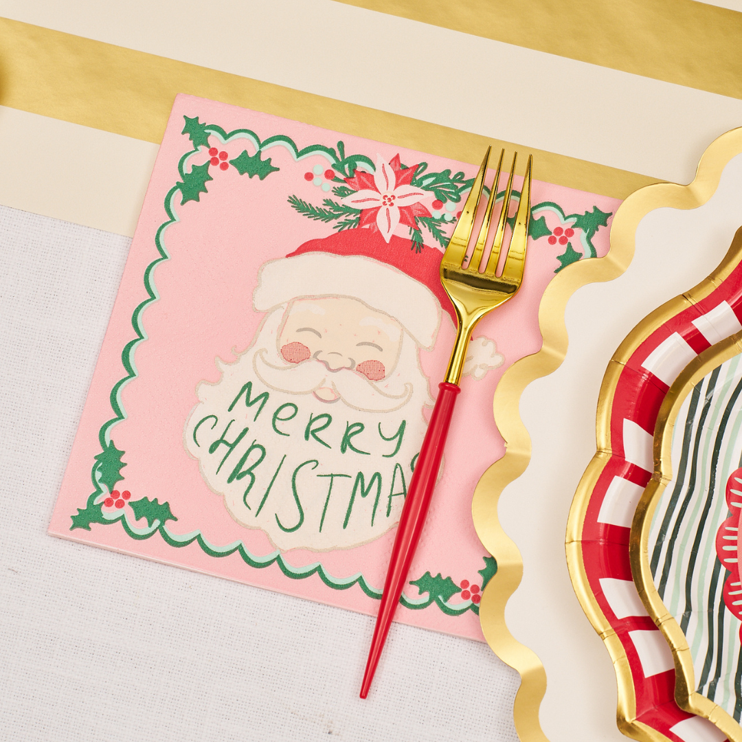A red and gold fork rests on a Sugarplum Tis the Season Lunch Napkin (20pk), featuring a Santa illustration and “Merry Christmas” text, set beside gold-rimmed festive paper tableware on a white and gold-striped tablecloth—perfect for holiday gatherings.