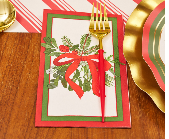 A gold fork and knife are arranged on a Sophistiplate Home for the Holidays Guest Towel napkin, featuring holiday greenery and a red bow design, set on a wooden table next to a gold-rimmed plate.