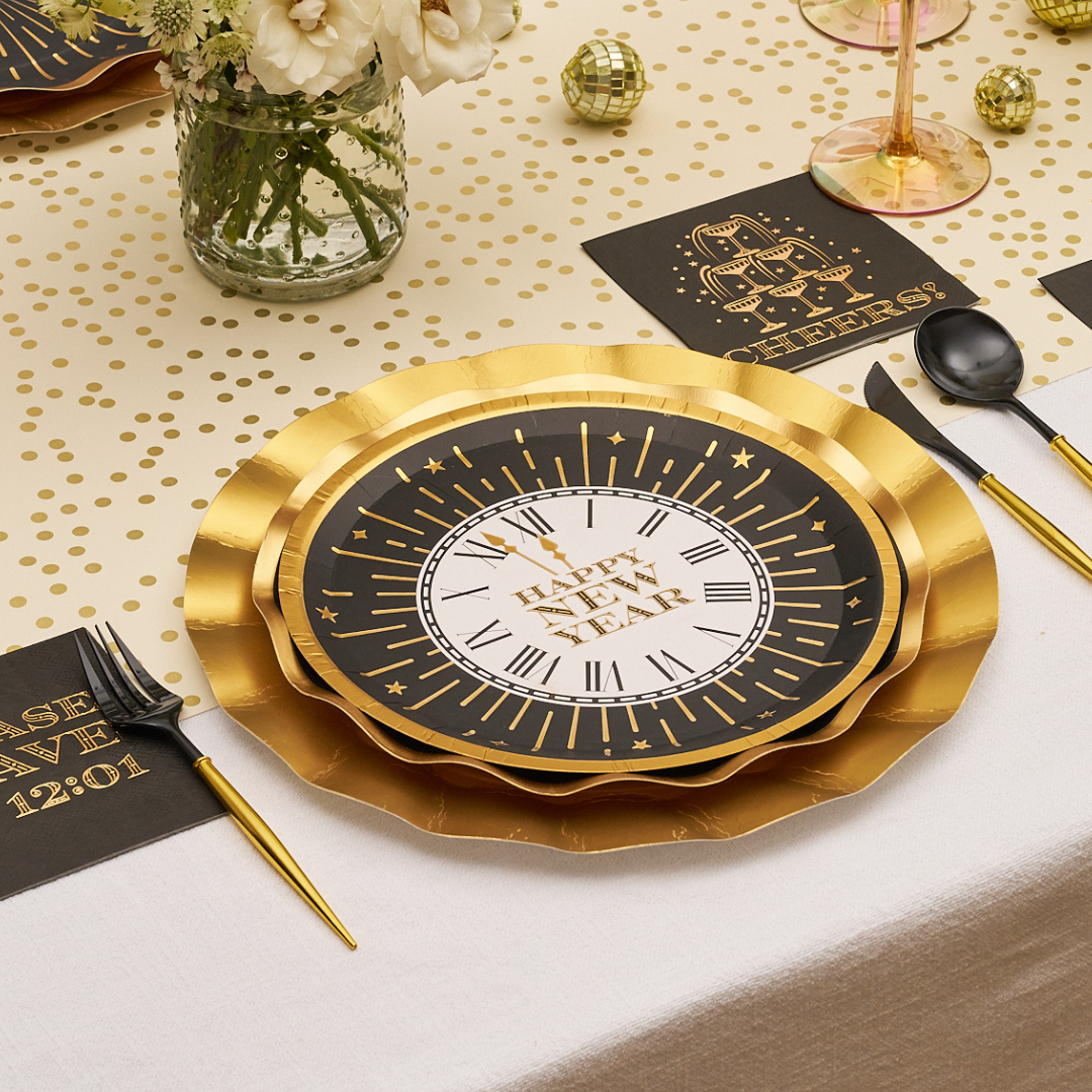 A round, black and white Midnight Sparkle Salad Plates/8pk featuring a clock face design that reads Happy New Year, layered on a larger scalloped-edge gold charger plate.